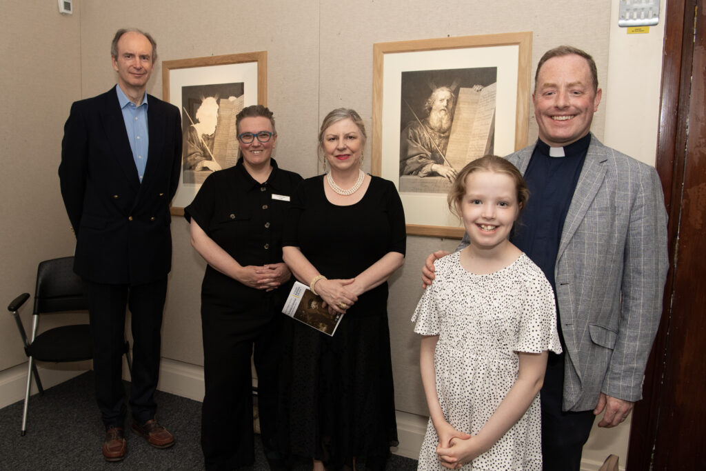 Five people standing in front of two prints on a beige coloured wall at exhibition launch in Armagh County Museum: Governor and Guardian Brett Hannam in black suit and blue shirt,; Archivist Thirza Mulder in black jumpsuit; Museum Mentor Noreen Cunningham in black dress and pearl necklass, and Governor and Guardian Peter Thompson in clerical dress, and miss Sarah Thompson in a flowery dress.