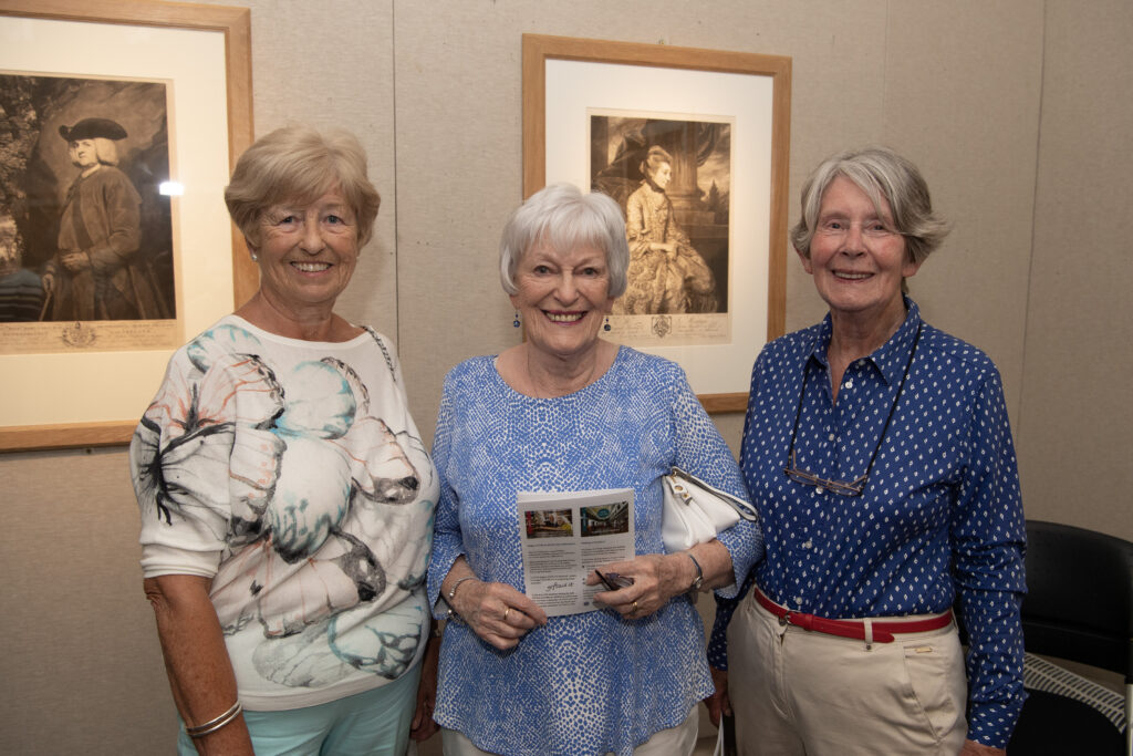 Three ladies standing in front of two prints on a beige coloured wall at exhibition launch in Armagh County Museum: Margaret Oakes in light blue trousers and white-blue flower print top, Ann Cassidy in blue printed top and Zena Dawson in cream trousers and blue white-spotted blouse.