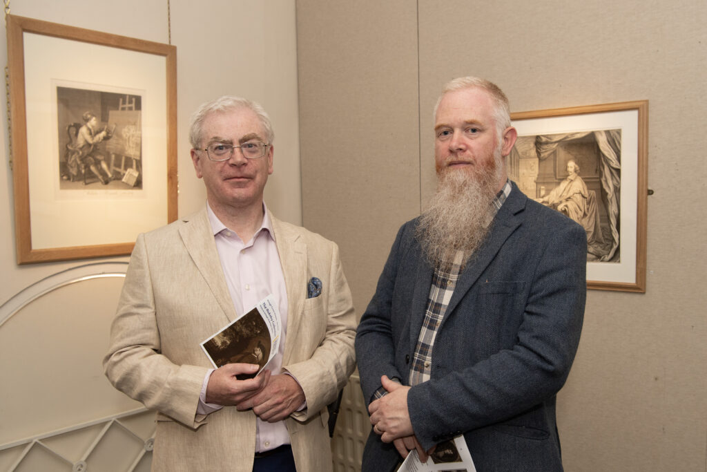 Two gentleman standing in front of two prints on a beige coloured wall at exhibition launch in Armagh County Museum: Curator of Newry and Mourne Museum Ken Abraham in light shirt and cream blazer, and Library supporter David Dunlop, sporting ginger-grey beard, in tartan shirt and blue-grey blazer.