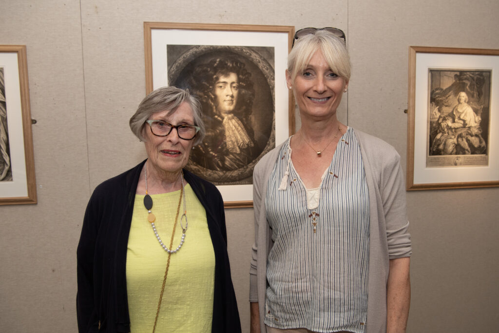 Two ladies standing in front of three prints on a beige coloured wall at exhibition launch in Armagh County Museum: Angela Boylan in yellow top and black cardigan and Lois Bennett in striped top and grey cardigan.