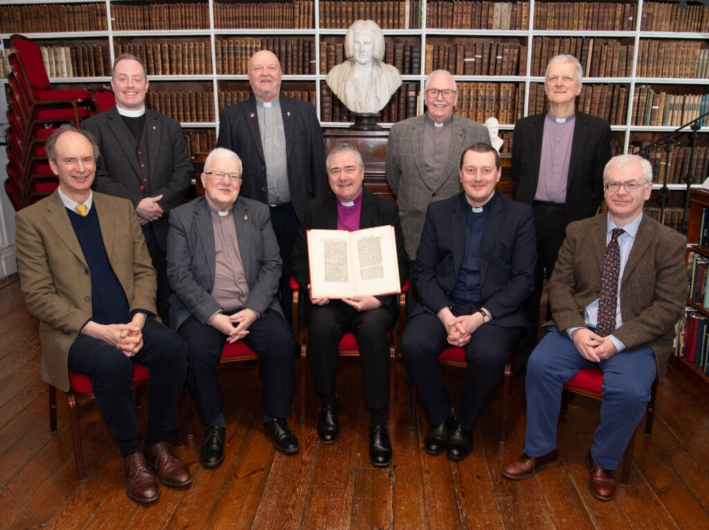 A group of gentlemen standing and seated in front of a buste of Archbishop Robinson in a historic library. The man in the centre is holding an copy of the 1773-1774 Act of Parliament.