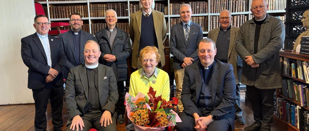Carol Conlin, seated, holding flowers, surrounded by the Governors and Guardians of Armagh Robinson Library in the Library's Long Room.
