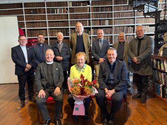 Carol Conlin, seated, holding flowers, surrounded by the Governors and Guardians of Armagh Robinson Library in the Library's Long Room.