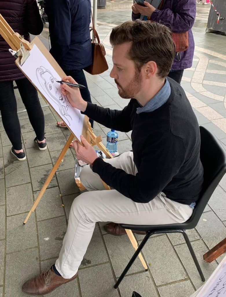 A man in khaki trousers and blakc sweater is sitting at an easel in a shopping centre, drawing a charicature.