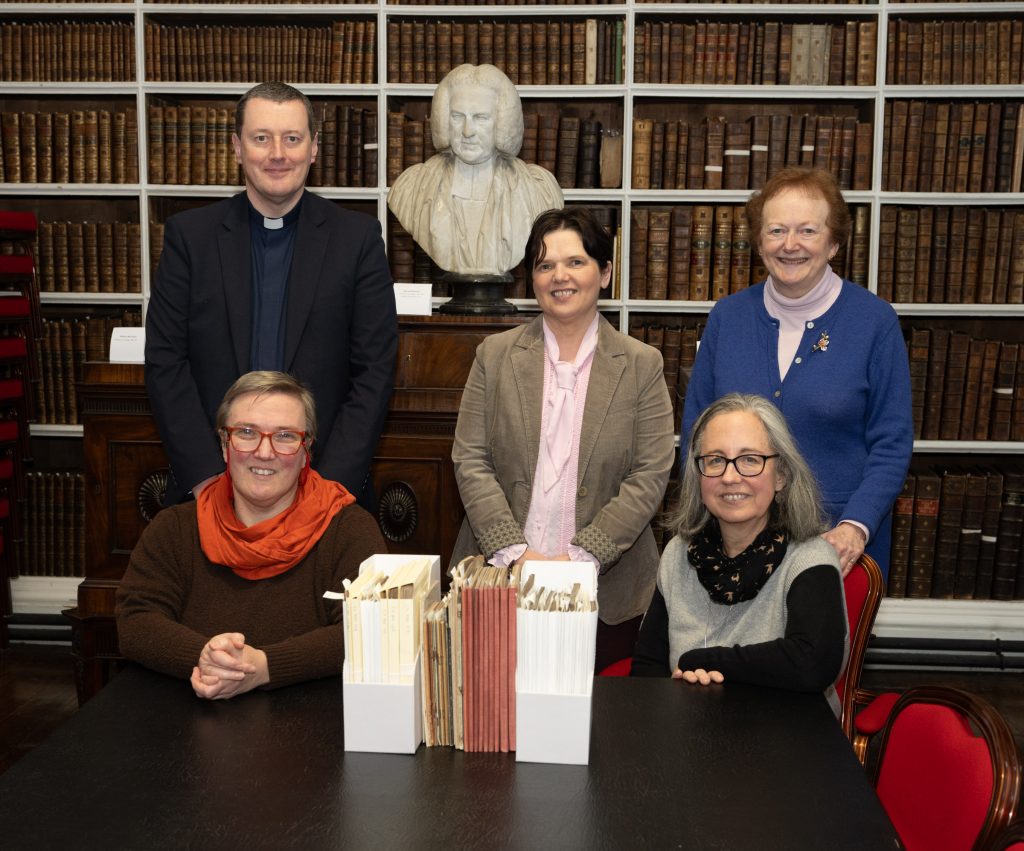 Visit NIMC Director Heather McGuicken. Five people standing and seated in front of the bust of Archbishop Robinson. In front of them is a table with part of the pamphlet collection in their old folders, and part re-packaged in archival-quality envelopes and boxes.