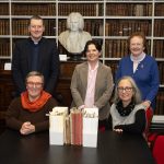 Five people standing and seated in front of the bust of Archbishop Robinson. In front of them is a table with part of the pamphlet collection in their old folders, and part re-packaged in archival-quality envelopes and boxes.