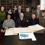 Ten people standing and seated behind a table in an historic library, with an open copy of the 1776 American Atlas in front of them