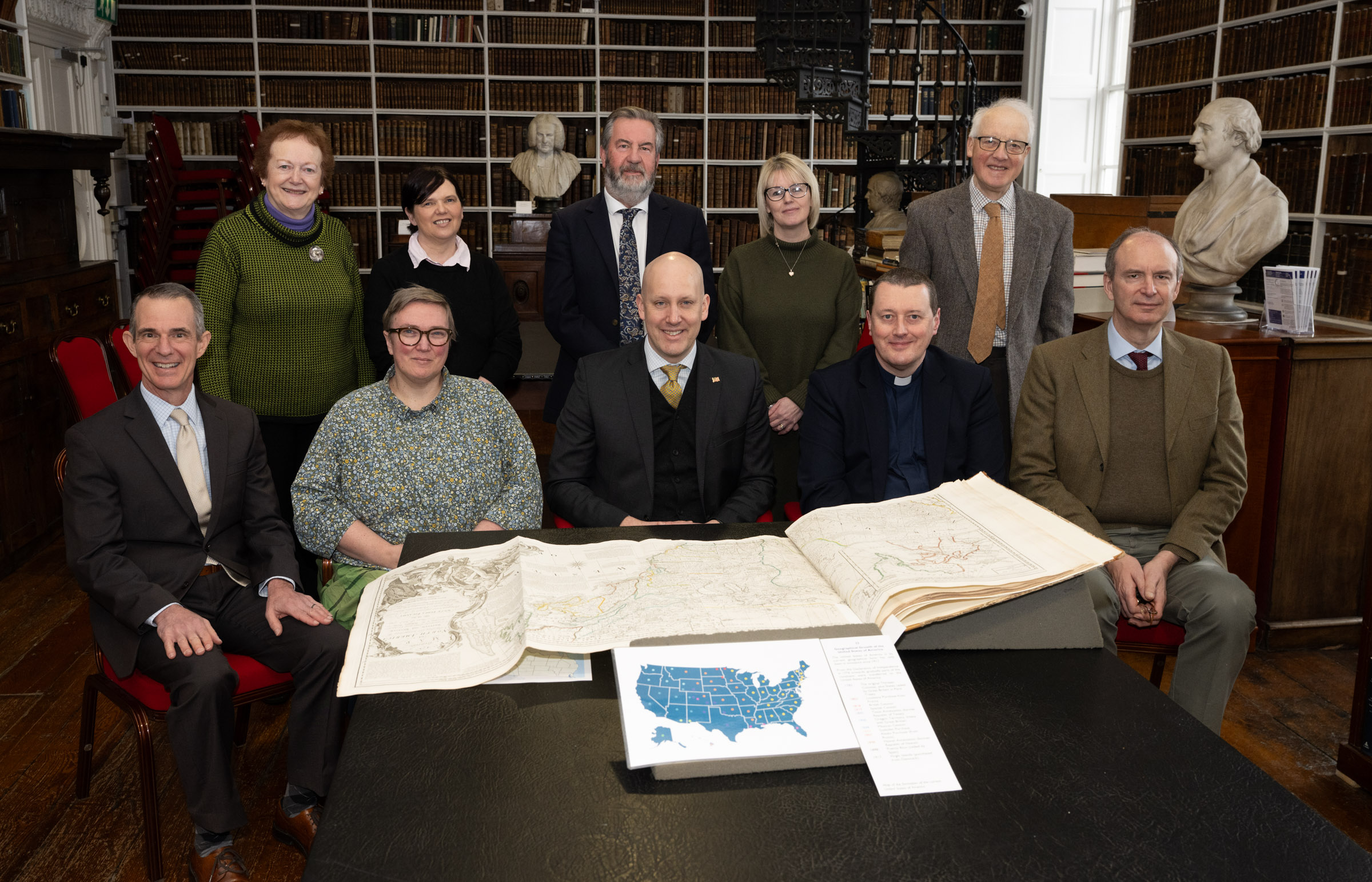 Ten people standing and seated behind a table in an historic library, with an open copy of the 1776 American Atlas in front of them